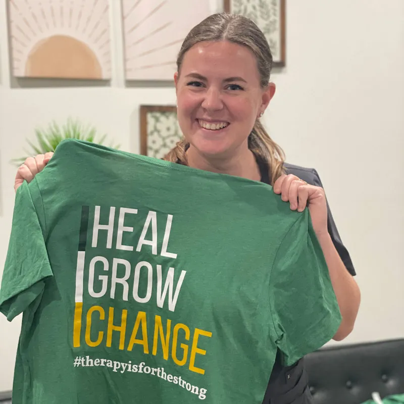 Smiling person holds a green shirt with the words "HEAL GROW CHANGE #therapyisforthestrong.