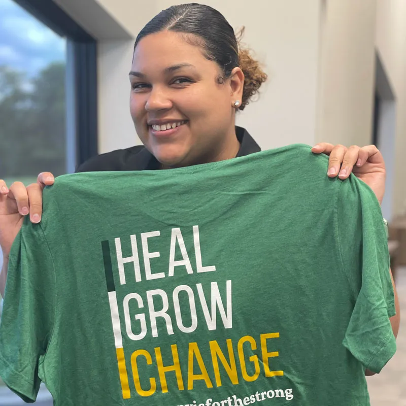 Person smiling and holding a green t-shirt with the words "Heal, Grow, Change.