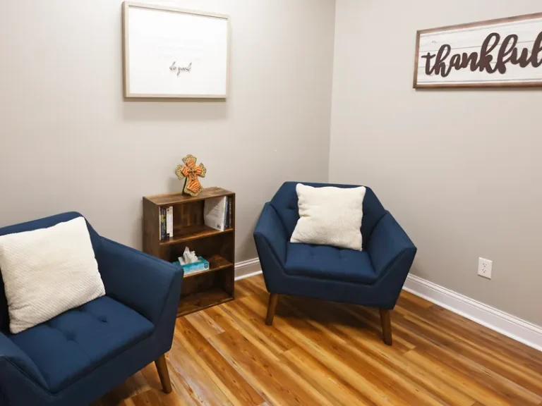Two blue chairs with white pillows face a small bookshelf in a cozy room with wooden floor and inspirational wall art.
