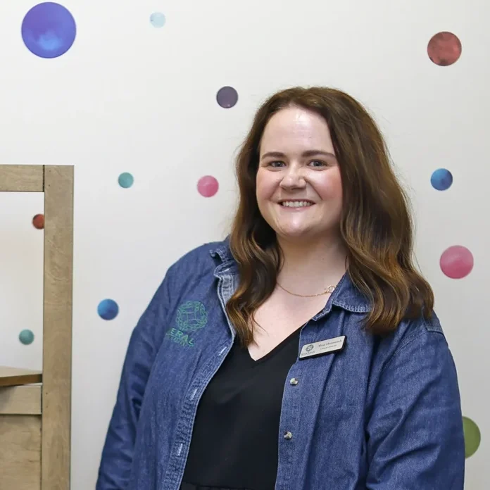 A woman stands by a colorful polka-dot wall and wooden shelf with playful decorations.