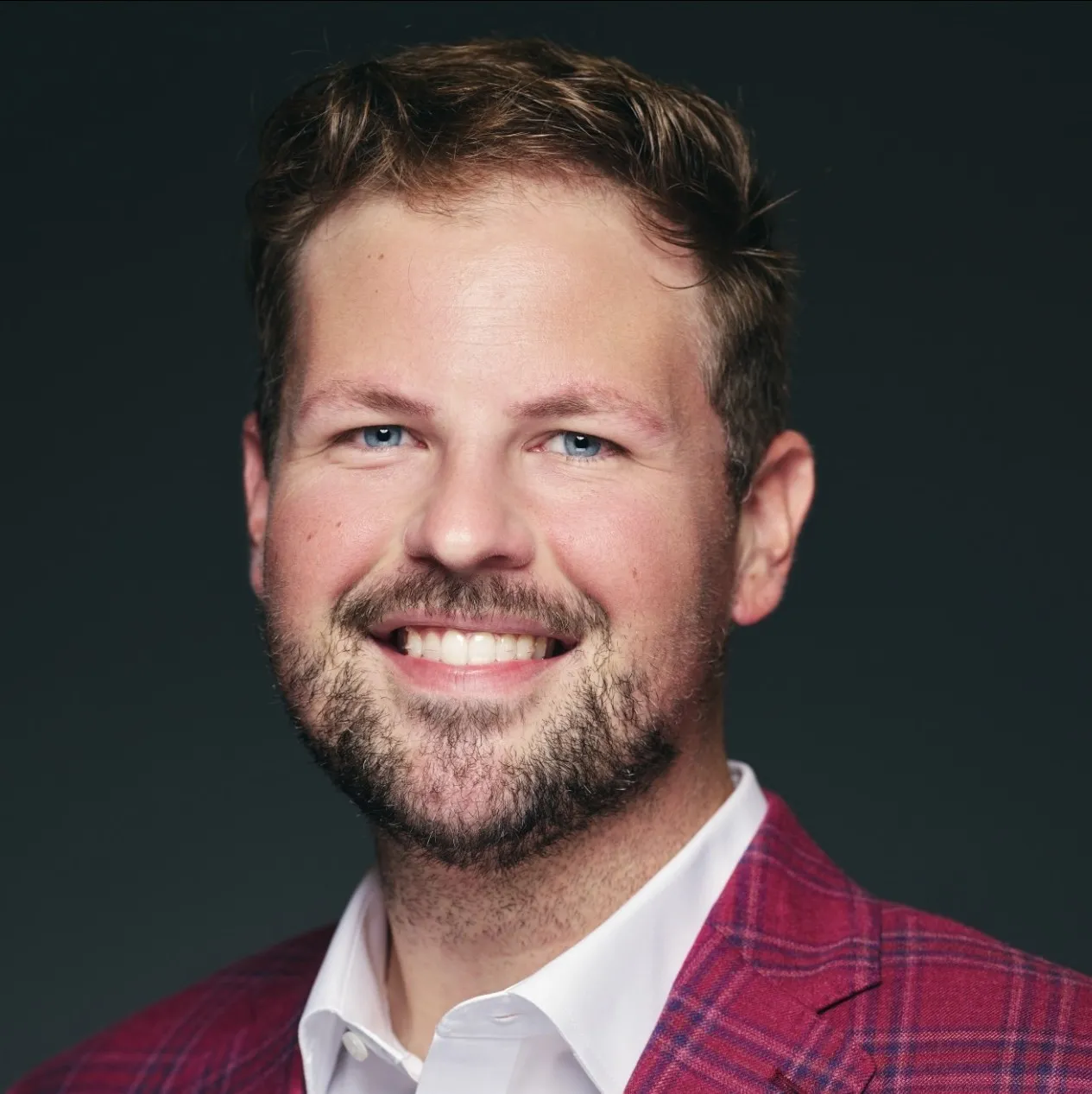 Smiling man in a red plaid jacket and white shirt with a dark background.