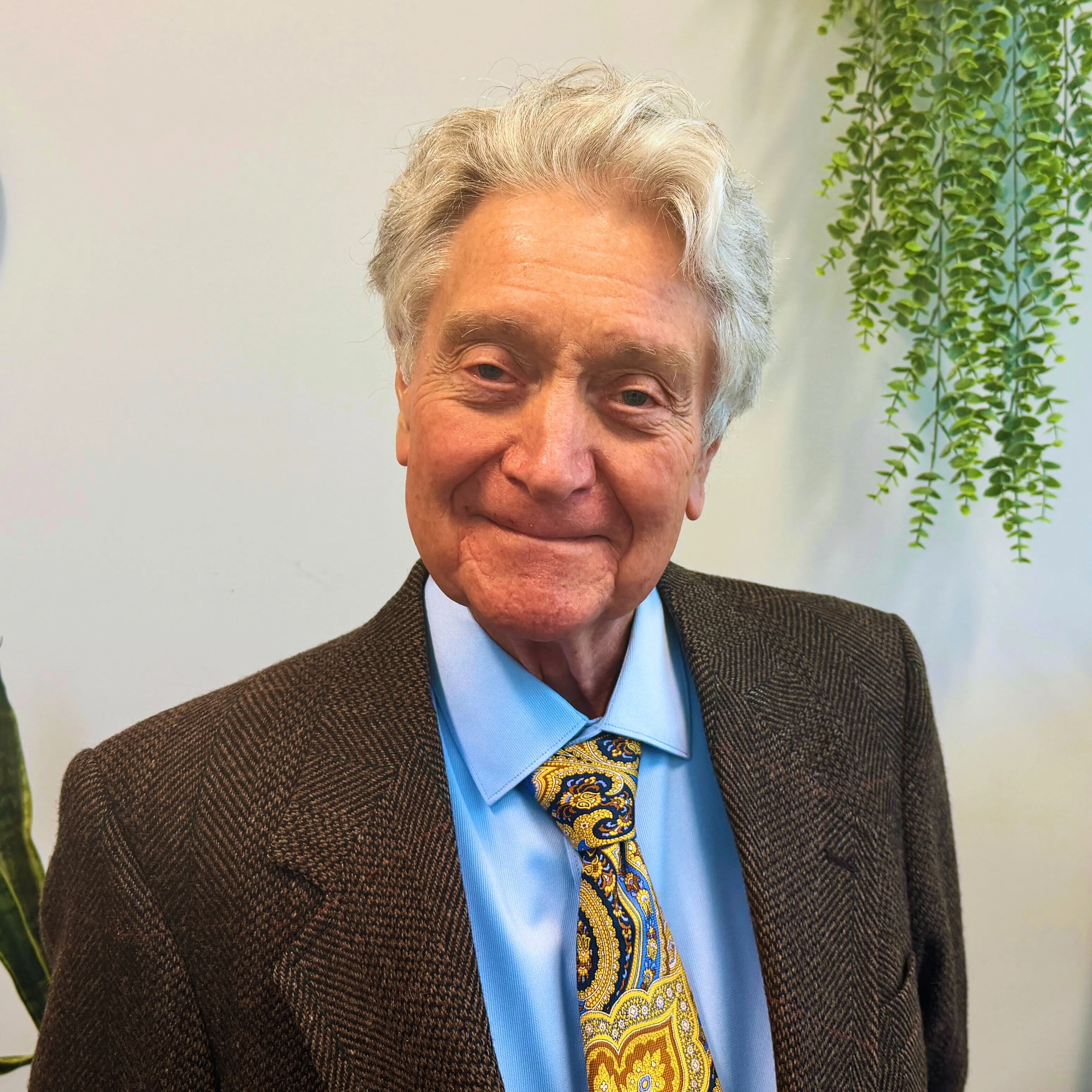 Elderly man in a suit and tie smiling in an office with plants and a clock.