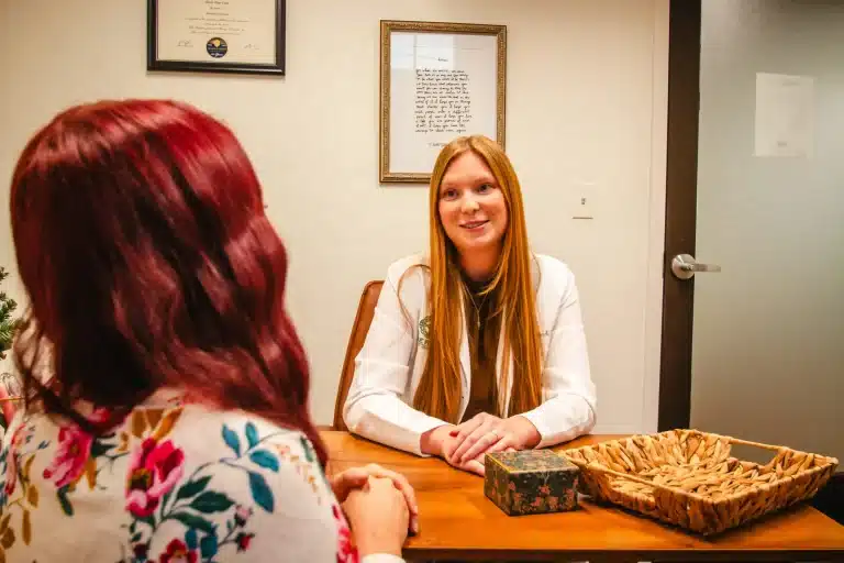 Two women are talking at a desk in an office setting.