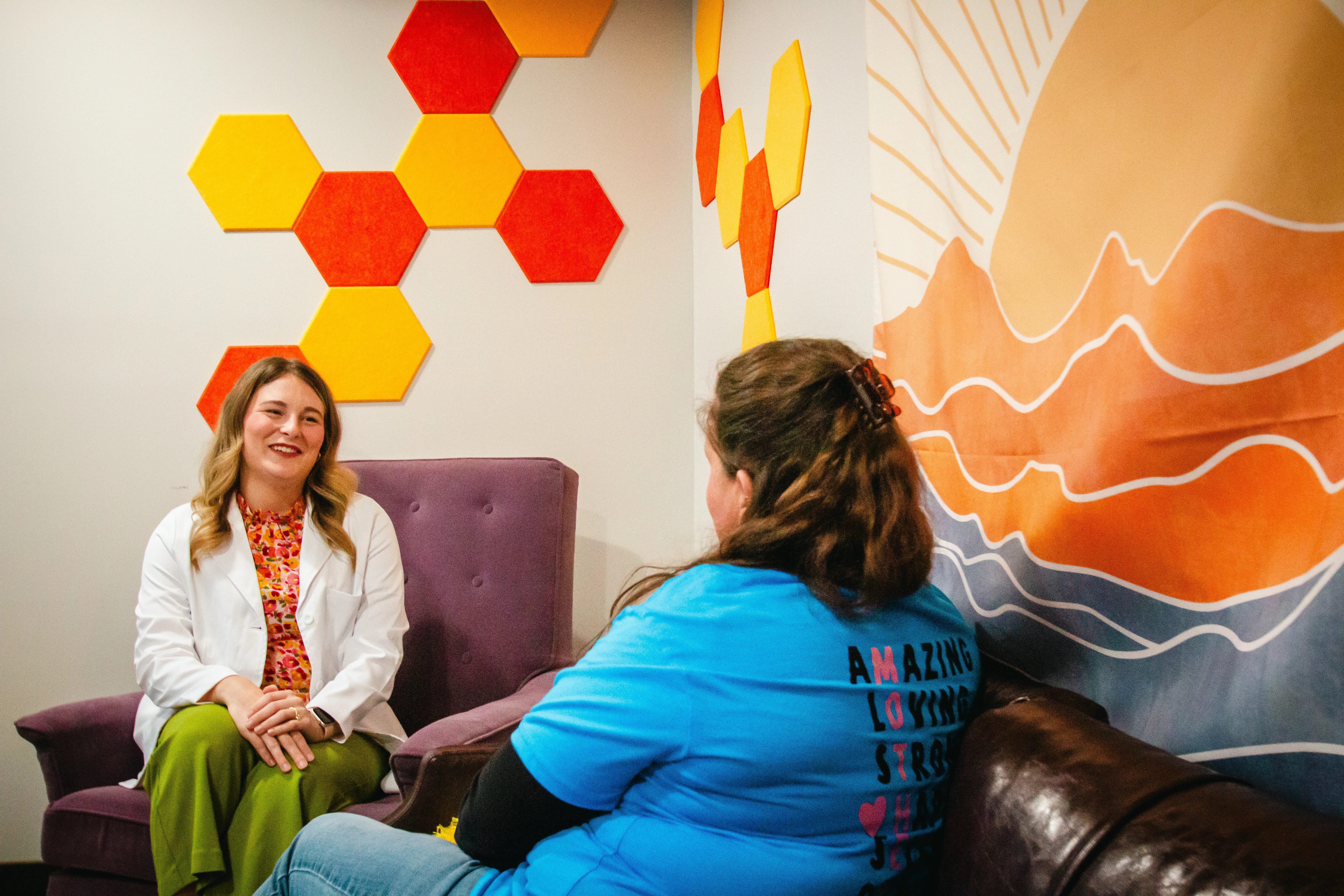 Two women talking in a room with colorful hexagon and landscape wall art.