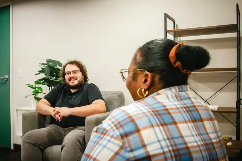 Two people are having a conversation in a cozy room with a plant and a shelf.