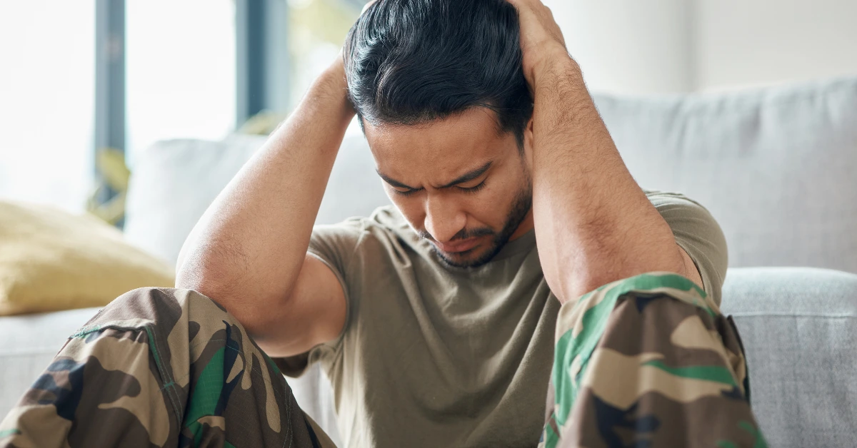 Veteran wearing camouflage pants sitting with hands on head, appearing stressed or overwhelmed while at home.