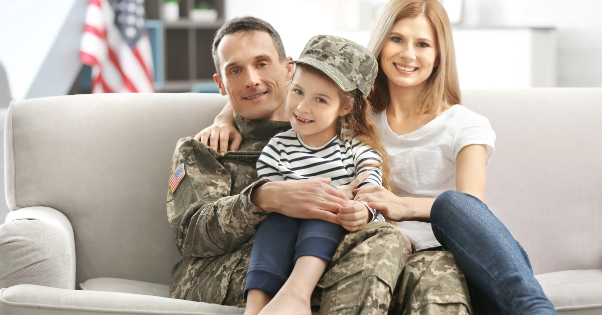 U.S. military veteran in uniform sitting on a couch with their spouse and young child, smiling together at home.