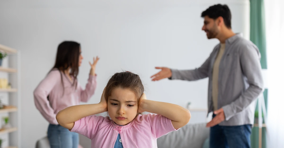Young child covering her ears while parents argue in the background, illustrating the emotional impact of childhood trauma and exposure to family conflict.