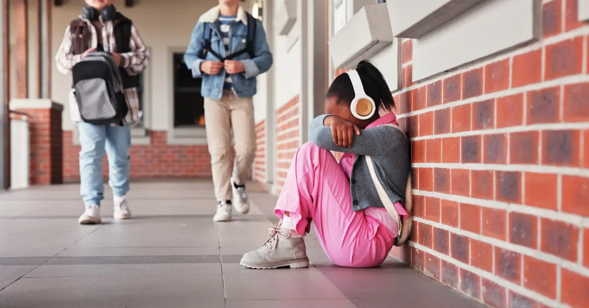 Child sitting alone outside school looking distressed while other students walk by, illustrating school anxiety in children.