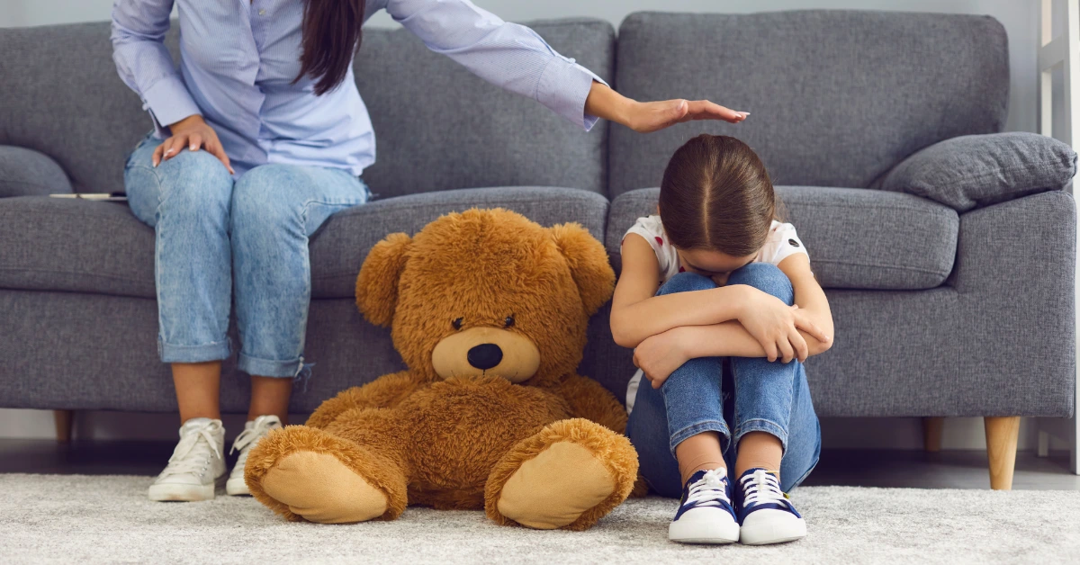 Young child sitting on the floor hugging her knees while a caregiver offers comfort, representing child grief counseling and emotional support after the loss of a loved one.