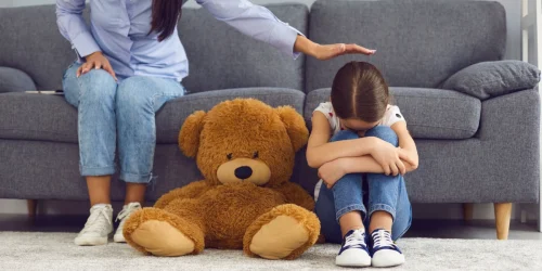 Young child sitting on the floor hugging her knees while a caregiver offers comfort, representing child grief counseling and emotional support after the loss of a loved one.