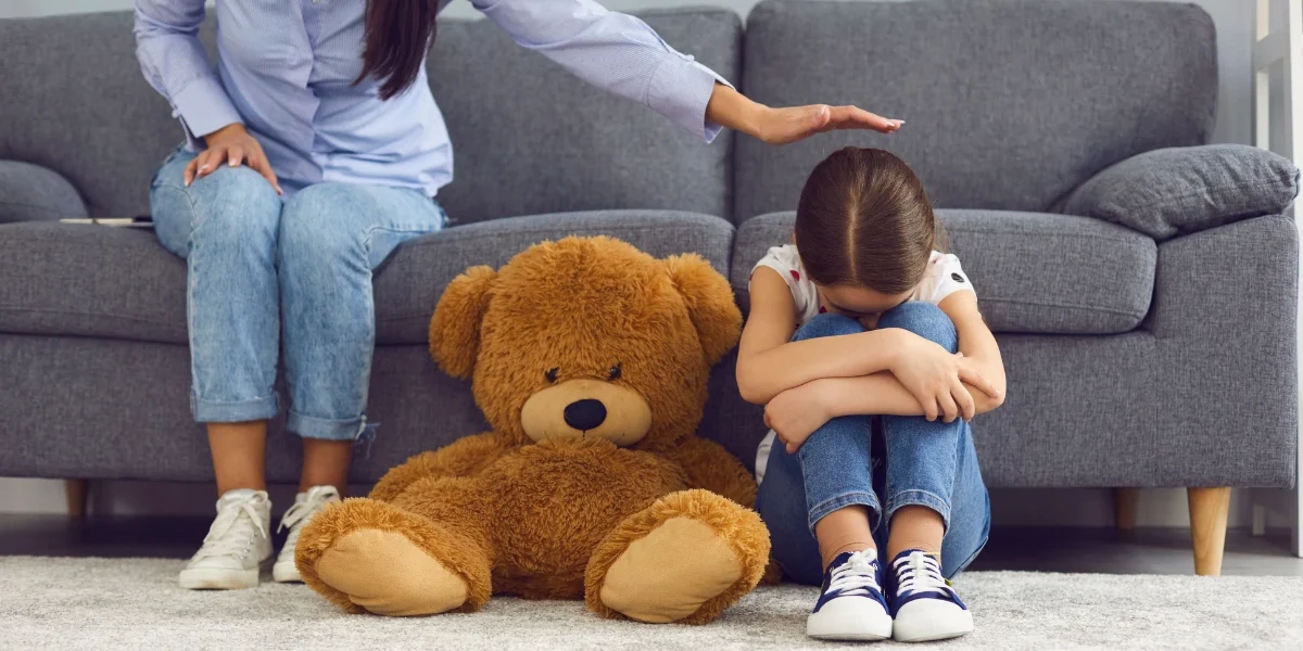 Young child sitting on the floor hugging her knees while a caregiver offers comfort, representing child grief counseling and emotional support after the loss of a loved one.