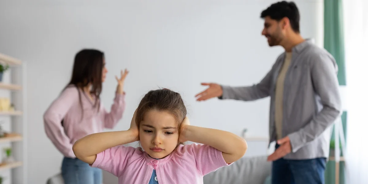 Young child covering her ears while parents argue in the background, illustrating the emotional impact of childhood trauma and exposure to family conflict.