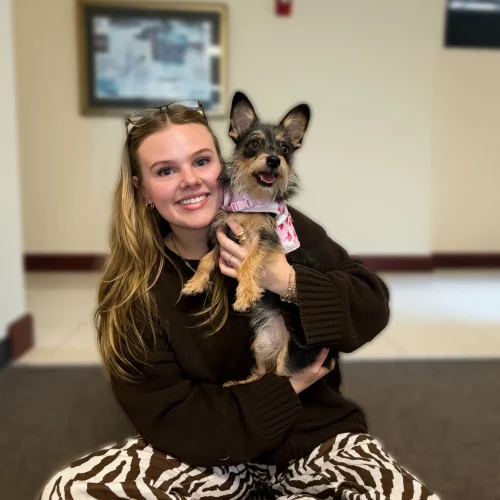 Young woman smiling while cuddling a small dog, representing the comfort, connection, and companionship animals can provide.