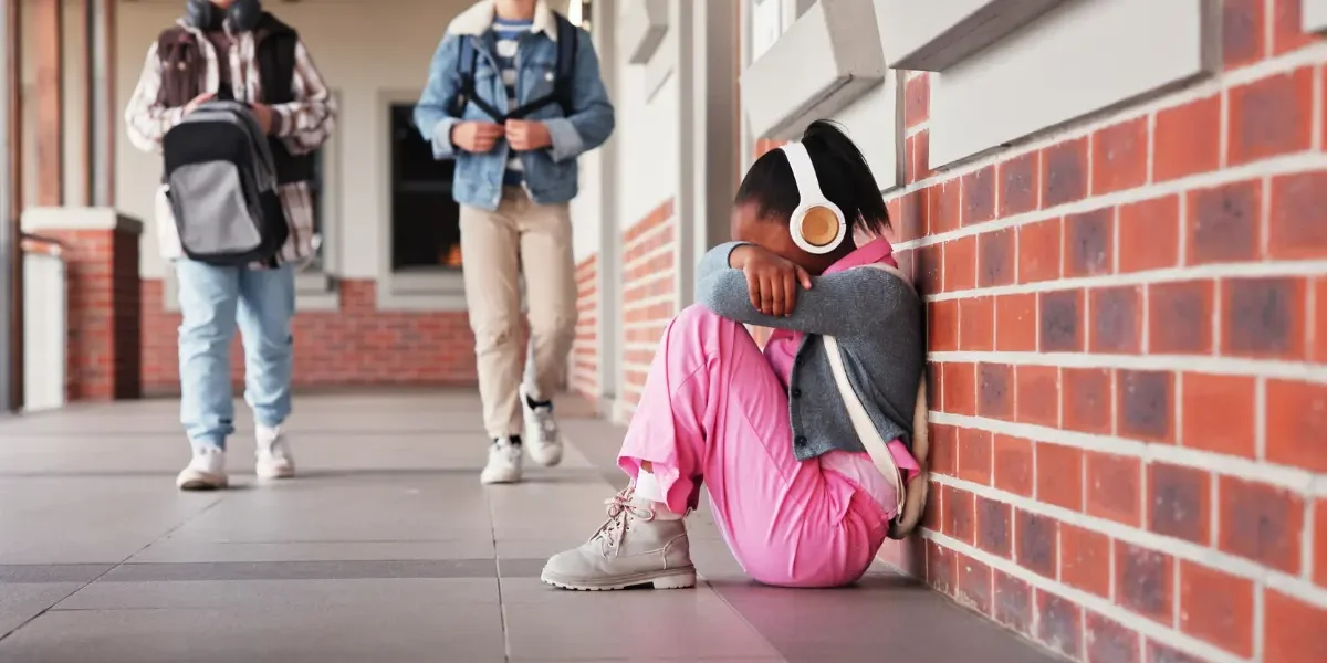 Child sitting alone outside school looking distressed while other students walk by, illustrating school anxiety in children.